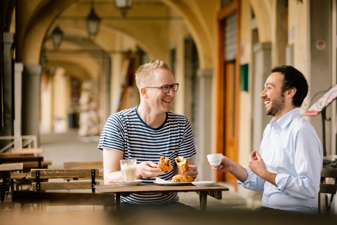 Jens Depenau lacht bei Espresso und Cornetto mit einem Freund unter alten Arkaden