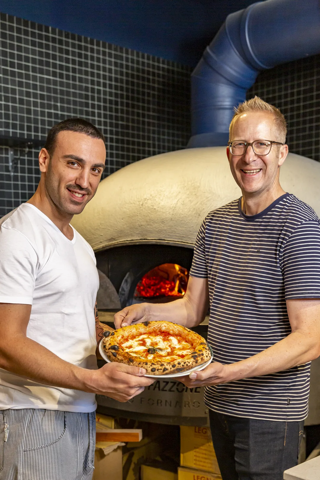 Jens Depenau und ein Pizzaiolo halten gemeinsam eine frisch gebackene Pizza vor dem Holzofen